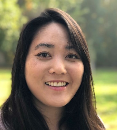 A smiling woman with long dark hair, looking at the camera outdoors with greenery in the background.